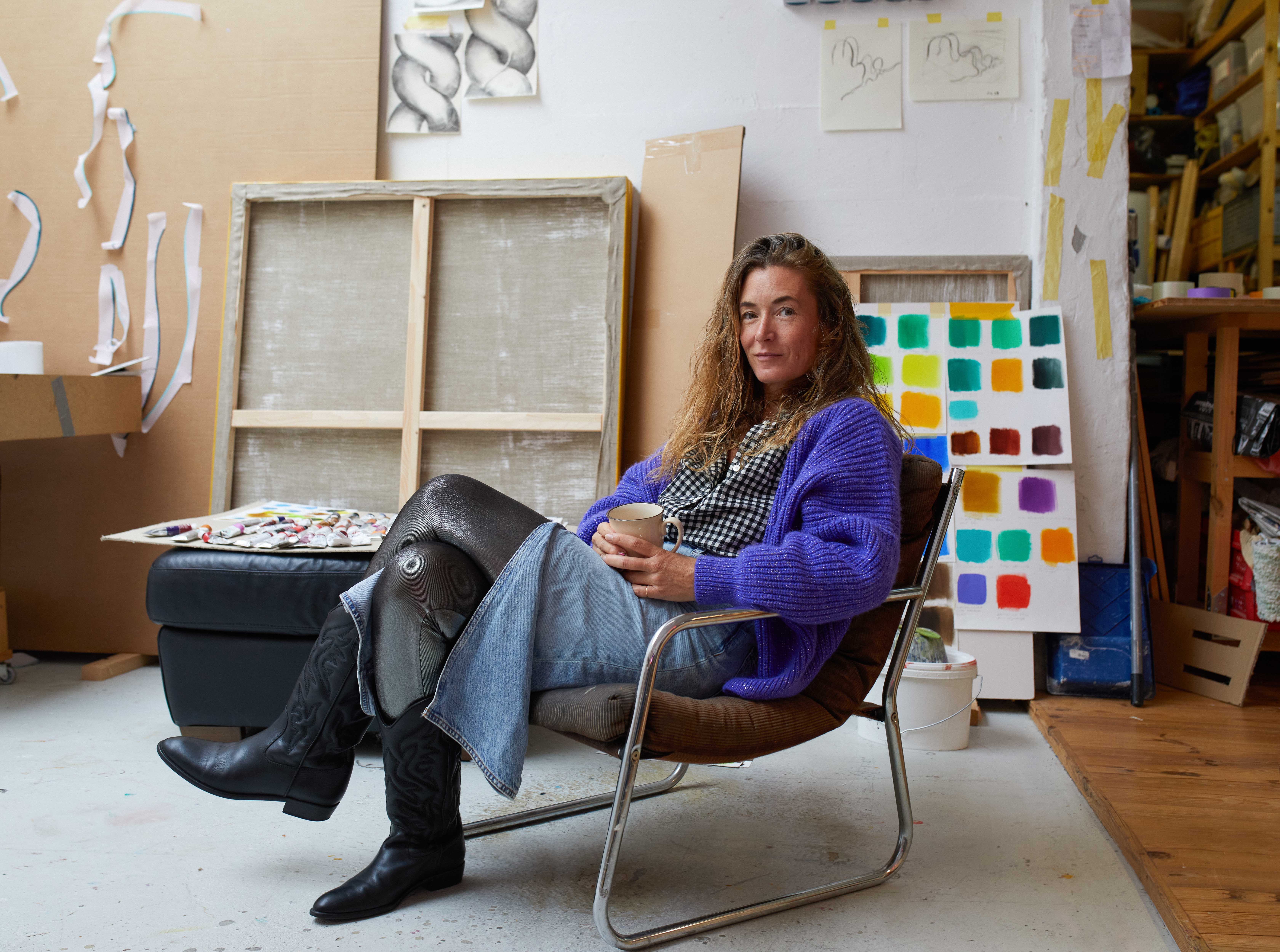 KI generiert: Eine Frau sitzt entspannt mit einer Tasse in der Hand in einem kreativen Studio. Um sie herum sind Leinwände, bunte Farbtafeln und Kunstmaterialien verteilt.