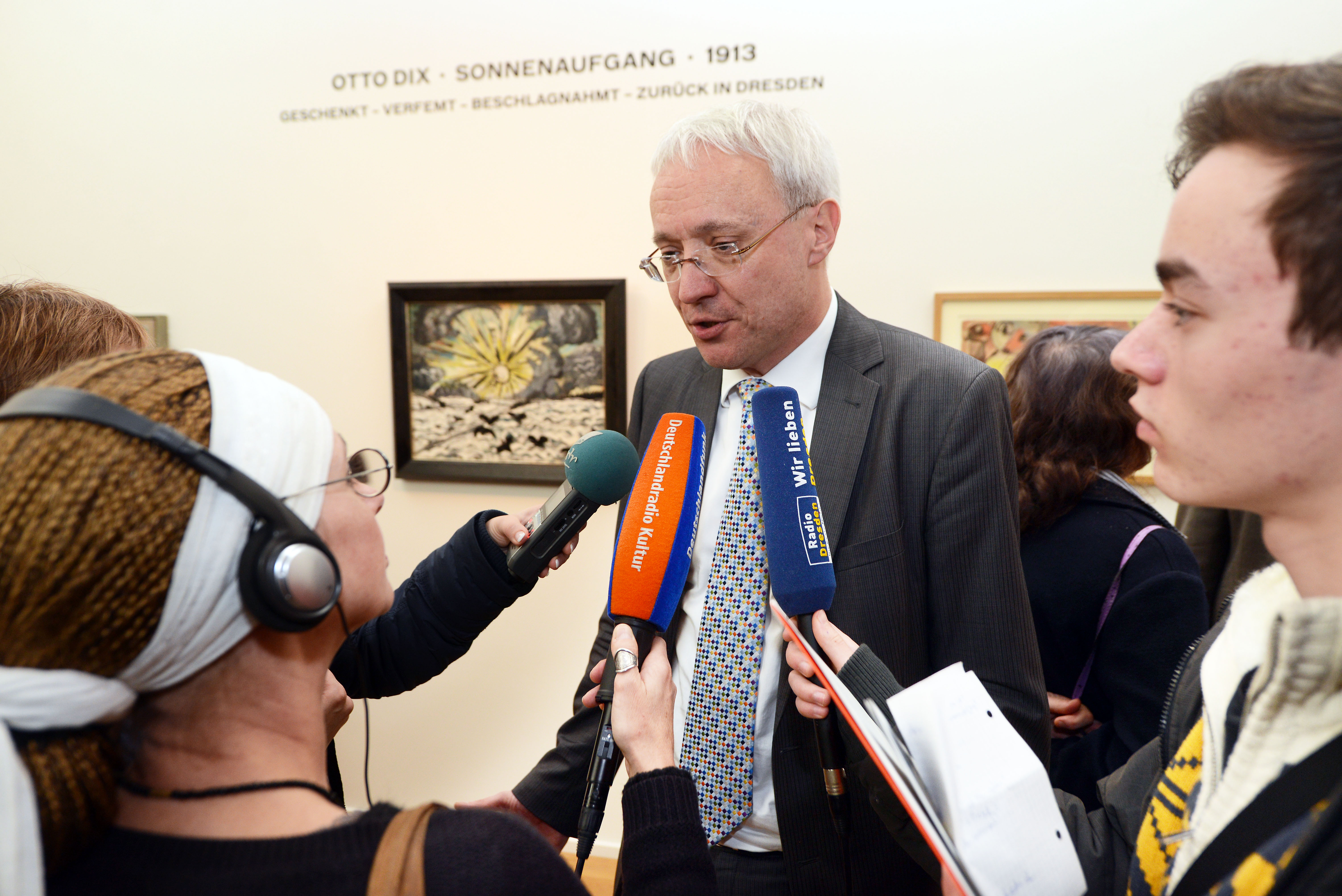 KI generiert: Ein Mann in Anzug wird von mehreren Journalisten mit Mikrofonen interviewt. Im Hintergrund sind Kunstwerke an der Wand zu sehen, darunter eines von Otto Dix.