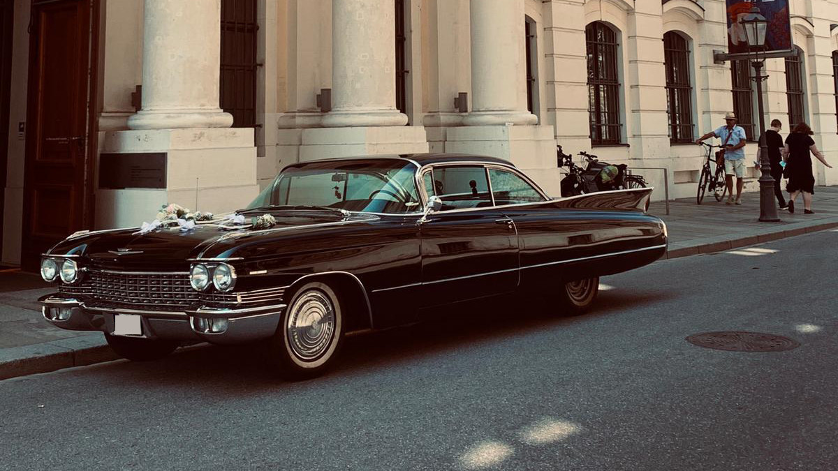 KI generiert: Das Bild zeigt ein klassisches schwarzes Auto, das vor einem historischen Gebäude mit großen Säulen geparkt ist. Auf der Motorhaube des Autos befinden sich Blumen, was auf eine Hochzeit oder Feierlichkeit hindeuten könnte.