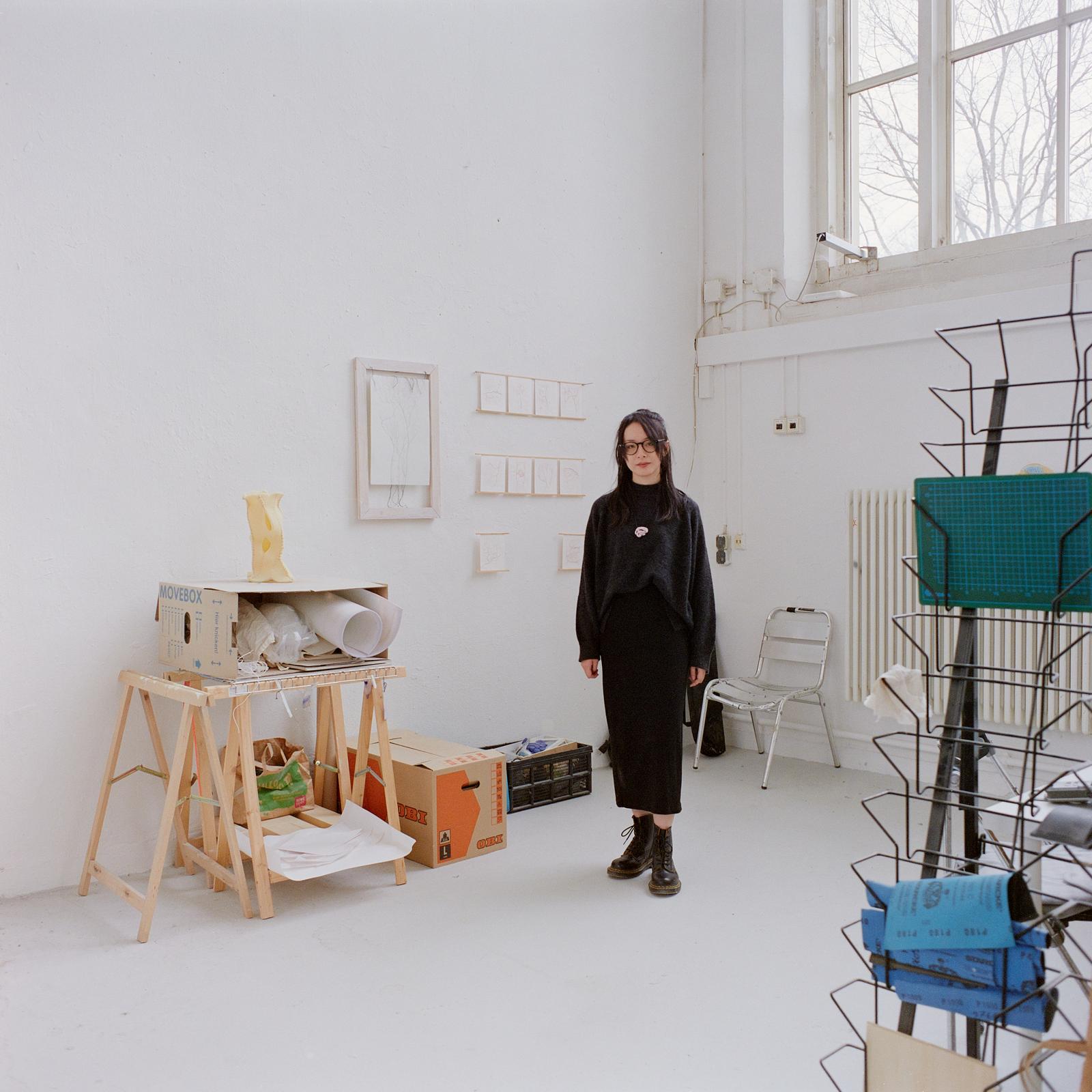 KI generiert: Das Bild zeigt eine Person in einem Atelier oder Arbeitsbereich mit weißen Wänden und großen Fenstern. Im Raum sind Kunstmaterialien und -werke zu sehen, sowie Stühle und verschiedene Utensilien.