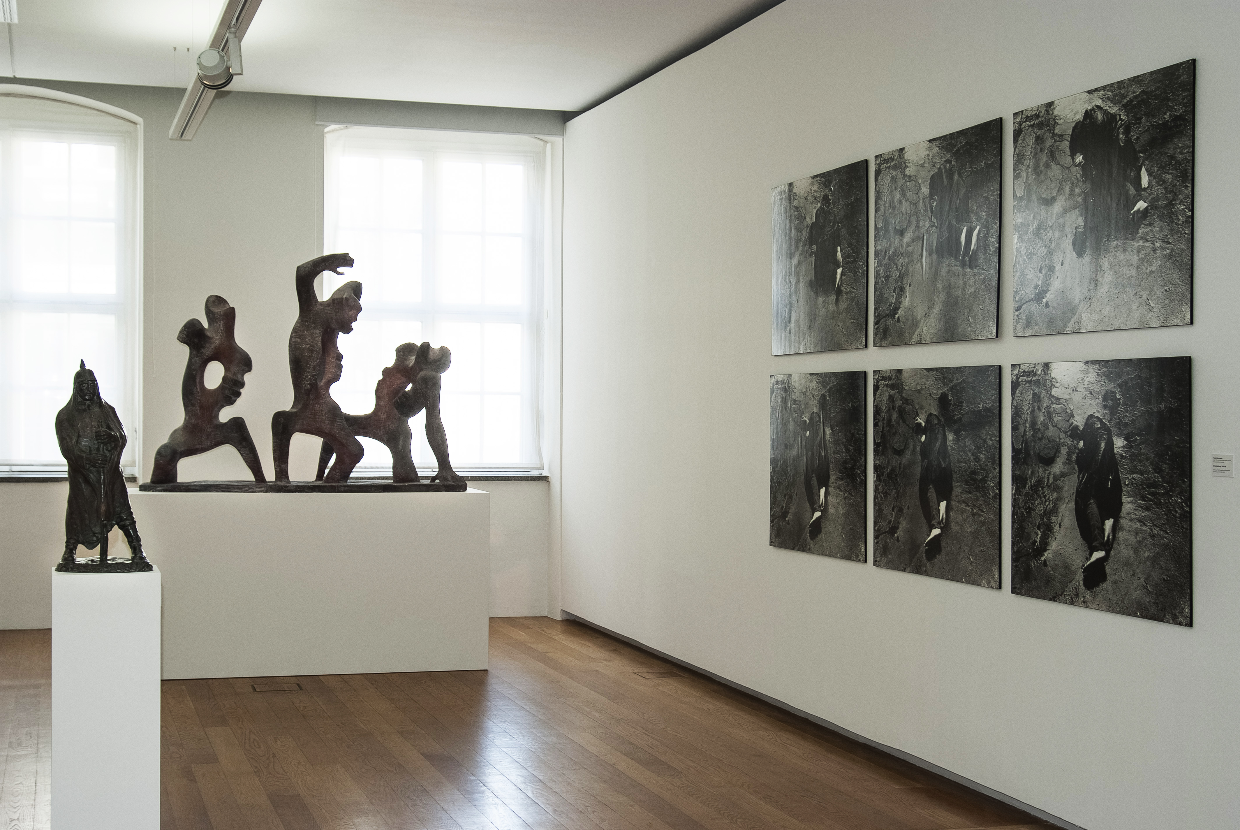 AI generated: The image shows an exhibition in an art-filled room in which several sculptures and six black and white photographs are presented on the wall. The sculptures are figurative and dynamic, while the photographs have a consistent theme.