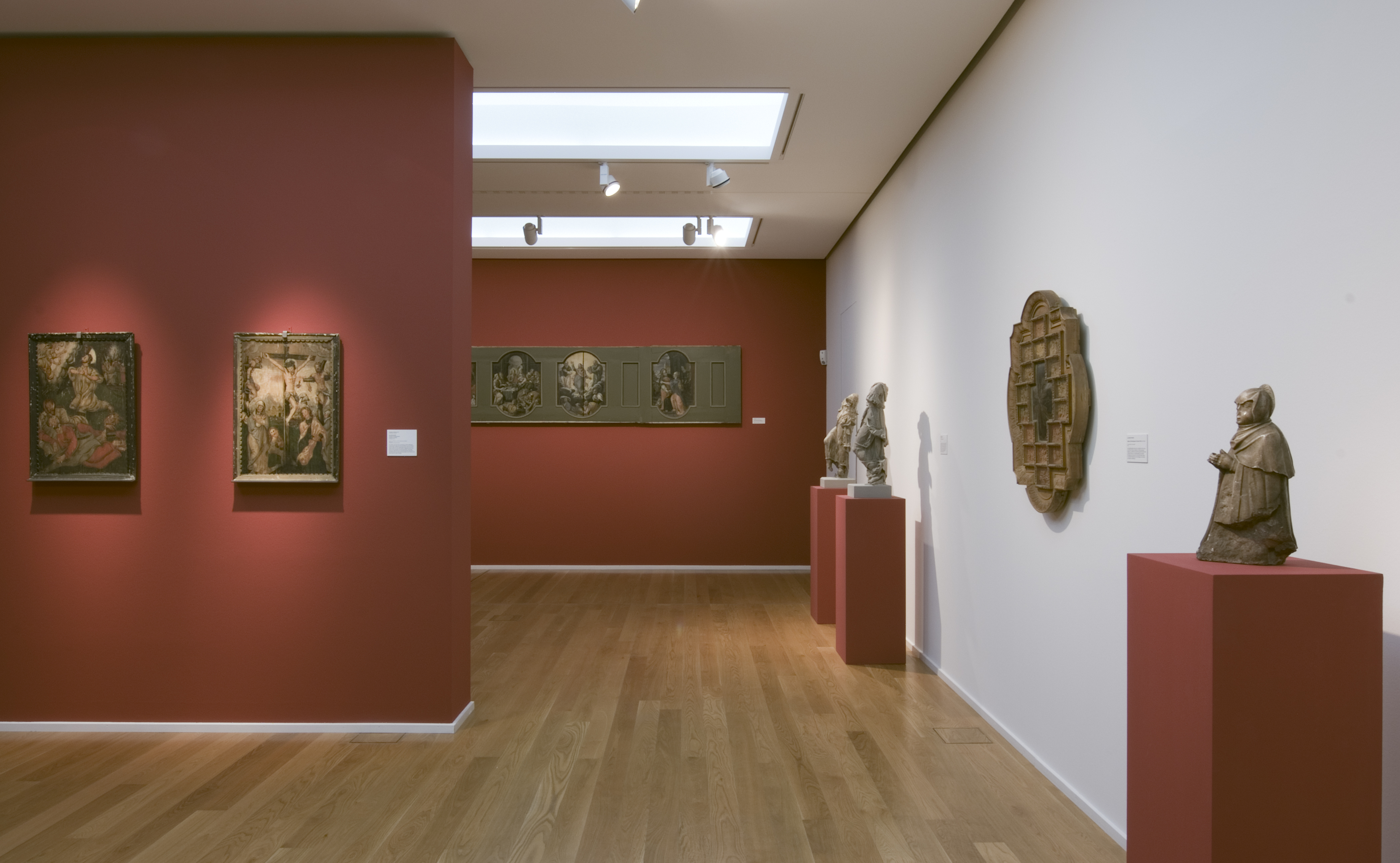 KI generiert: Das Bild zeigt eine Galeriewand in einem Museum, auf der Kunstwerke, darunter Gemälde und Skulpturen, auf roten Podesten oder an der Wand ausgestellt sind. Der Raum hat einen Holzfußboden und wird von oben beleuchtet.