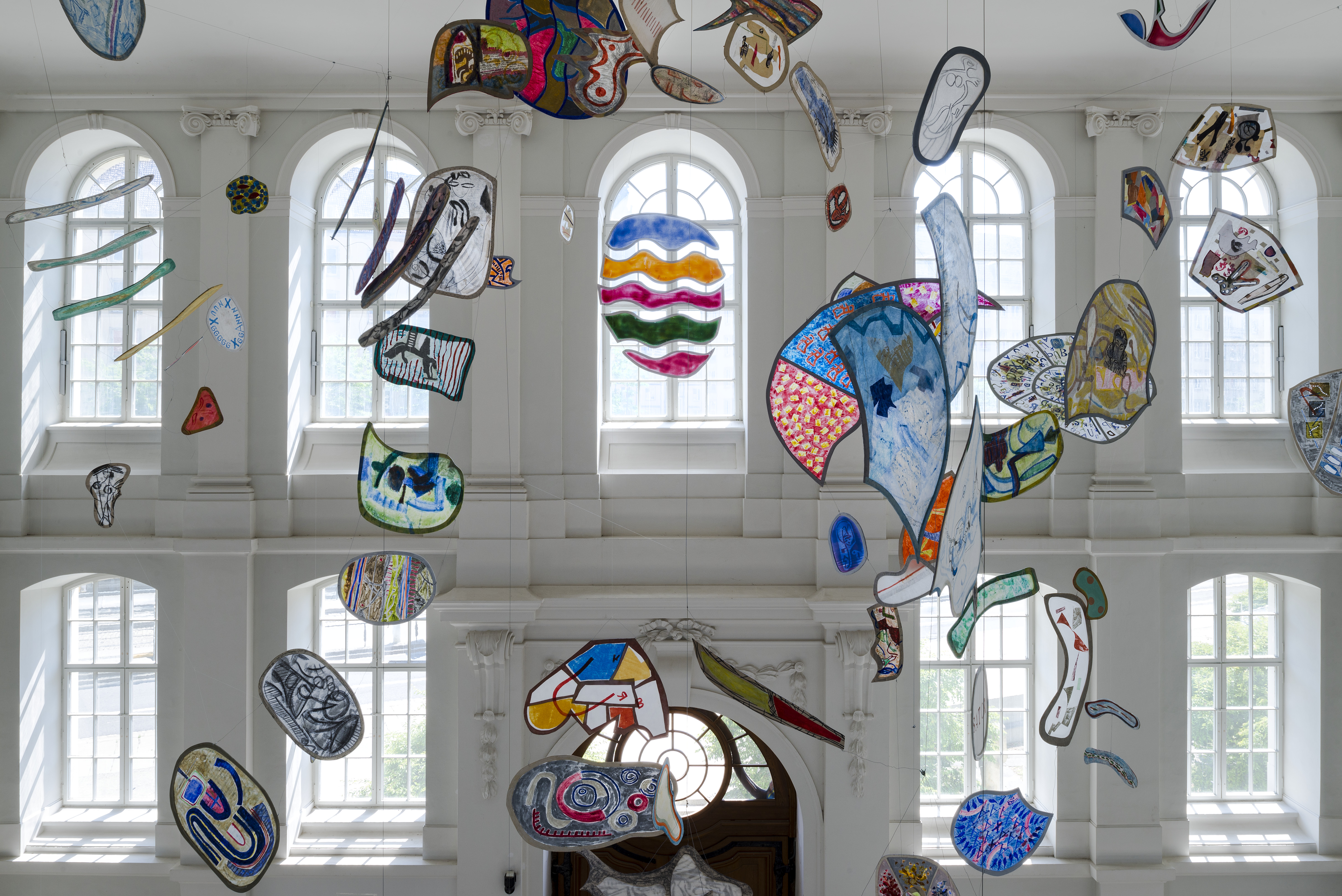 KI generiert: Das Bild zeigt eine Sammlung bunter, abstrakter Kunstwerke, die an der Decke eines großen, lichtdurchfluteten Raumes mit hohen Fenstern aufgehängt sind. Die schwebenden Formen und Farben schaffen eine lebendige, dynamische Atmosphäre.